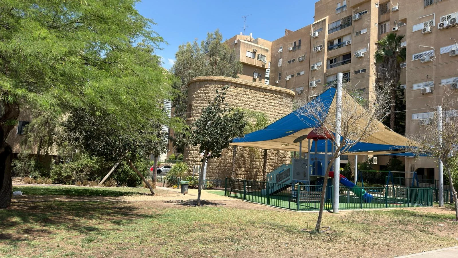 גינת משחקים לילדים עם מתקנים צבעוניים, מוצלת על ידי יריעת בד כחולה, לצד בנייני מגורים