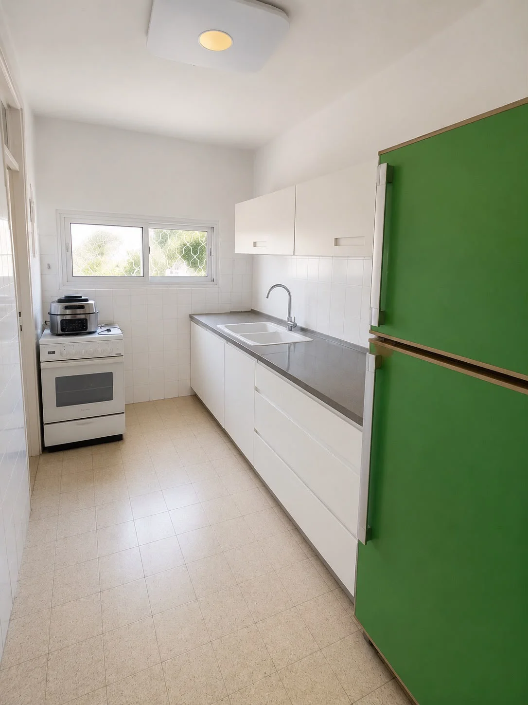 מטבח לבן עם ארונות עליונים ותחתונים, כיור כפול, כיריים לבנות ומקרר ירוק בולט. חלון קטן משקיף החוצה.