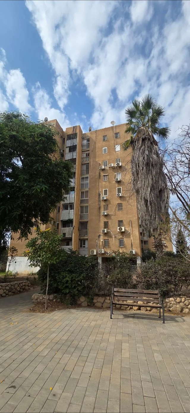 בניין מגורים בן שמונה קומות בבאר שבע, עם עצים וספסל בחזית.